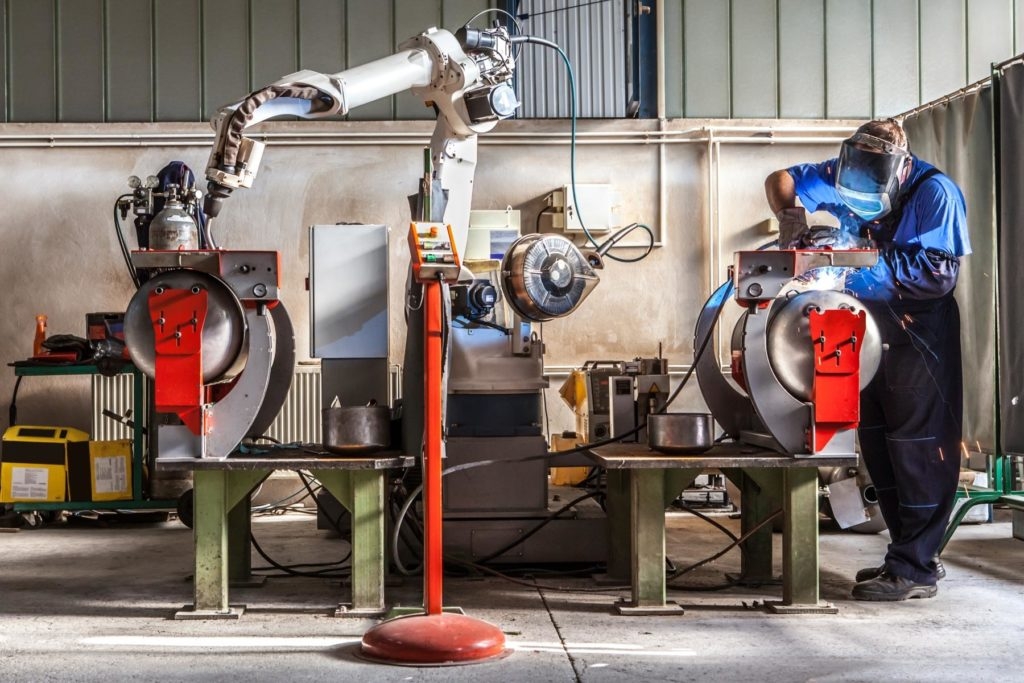 Man and robotic machine work together inside industrial building. The mechanical arm performs welds on metal components assisted by a worker who in turn manages welds manually.